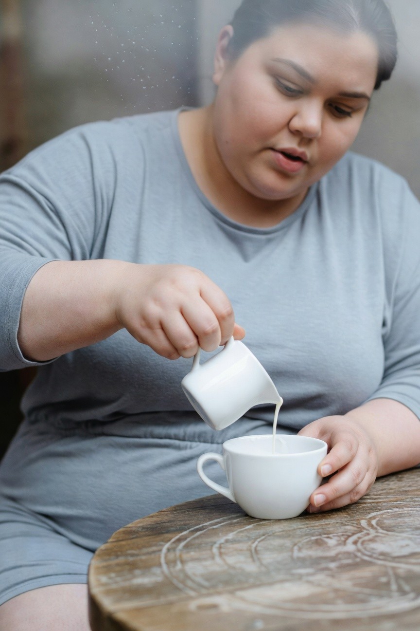 Plus-size woman in light gray long-sleeve romper pouring milk from creamer into cup at wooden table
