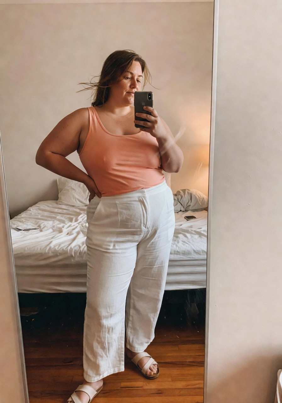 Plus-size woman taking a mirror selfie in a bedroom, wearing a sleeveless peach tank top, wide-leg white linen pants, and tan flat sandals, smiling softly while holding her phone
