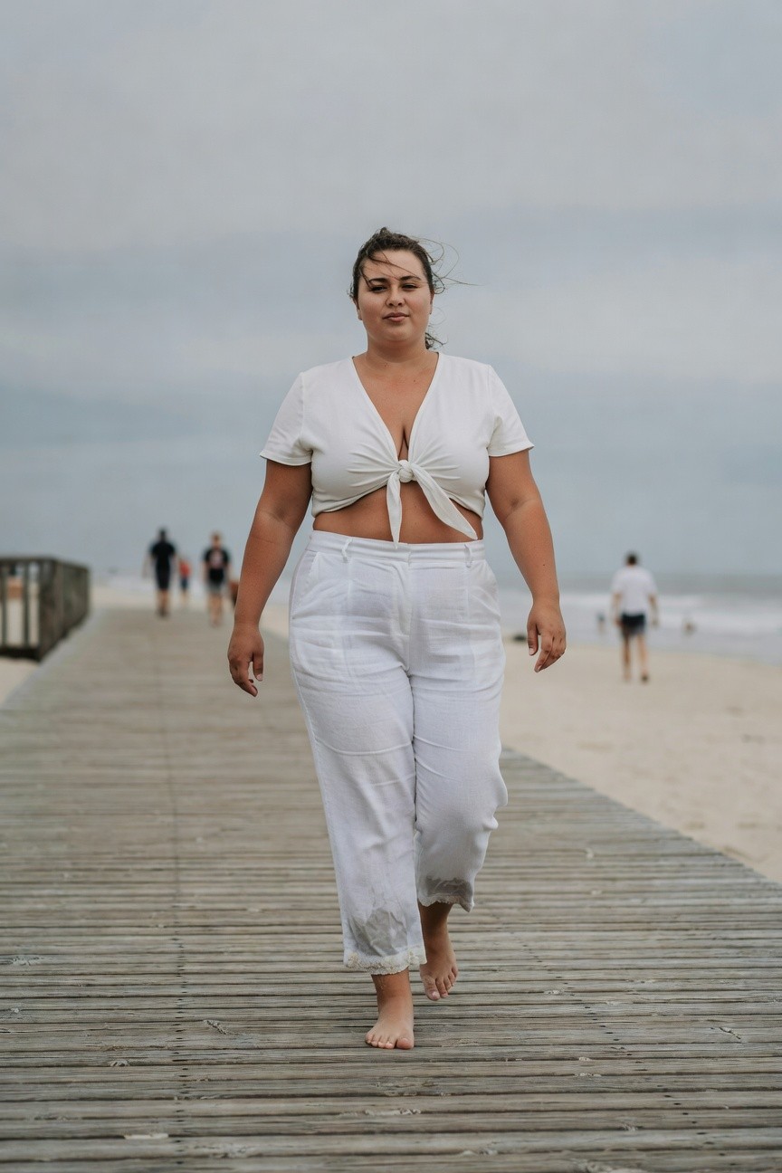 Plus-size woman walking barefoot in a white short-sleeved V-neck crop top tied at the front and matching wide-leg white pants on a wooden pier with beach in background