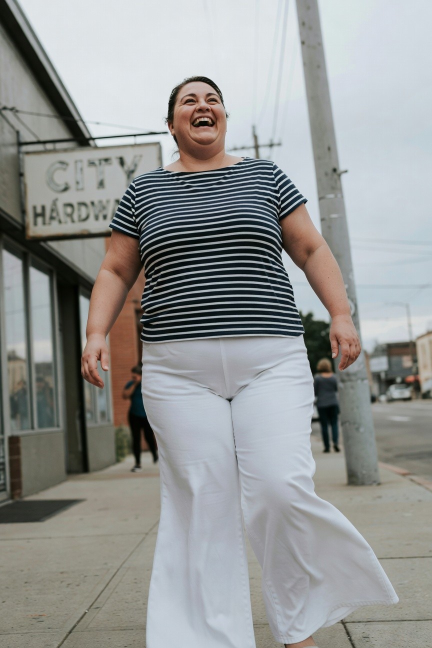 Plus size woman laughing while walking in navy and white horizontal striped short sleeve top tucked loosely into high waisted wide leg white pants, casual summer outfit on urban sidewalk