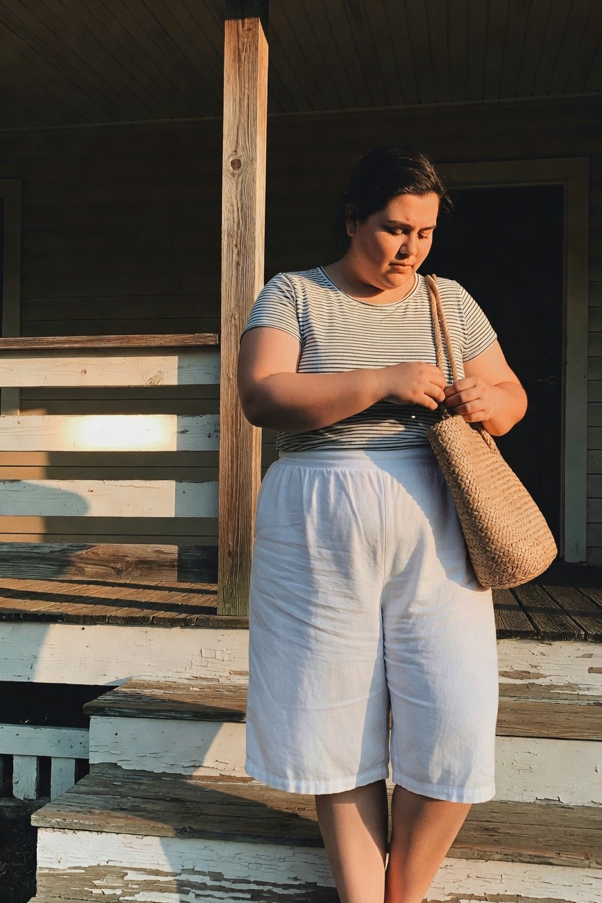 Plus-size woman with dark hair wearing a gray-and-white horizontal striped short-sleeve tee tucked loosely into high-waisted white linen wide-leg cropped pants, holding a large tan woven straw tote bag, standing on weathered wooden porch steps