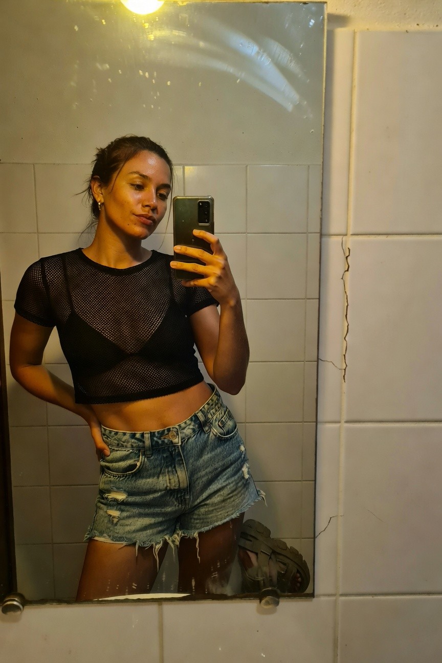 Woman taking a mirror selfie in a bathroom, wearing a sheer black mesh cropped top over a bralette, high-waisted light blue ripped denim shorts, and strappy sandals, with hair loosely tied back and small hoop earrings.