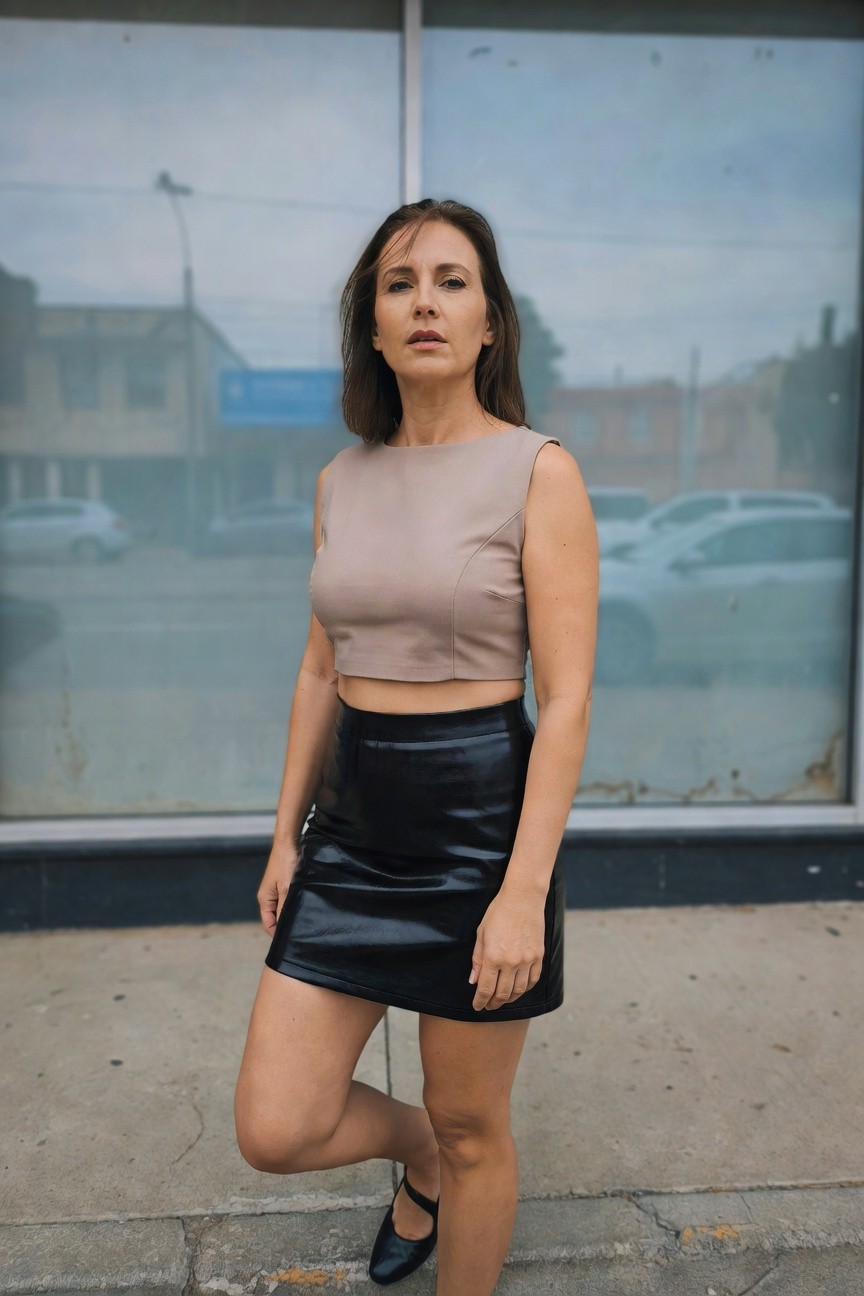 Woman in her 40s with shoulder-length brown hair standing sideways outdoors in front of a glass storefront, wearing a sleeveless beige cropped top exposing midriff, glossy black leather mini skirt, and black Mary Jane flats with ankle strap, one leg bent back against the wall