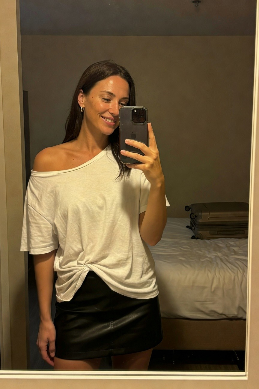 Mirror selfie of a woman smiling while holding her phone, wearing an off-shoulder white cotton t-shirt twisted at the waist tucked into a short black leather skirt, with hoop earrings and suitcases visible in a hotel room background