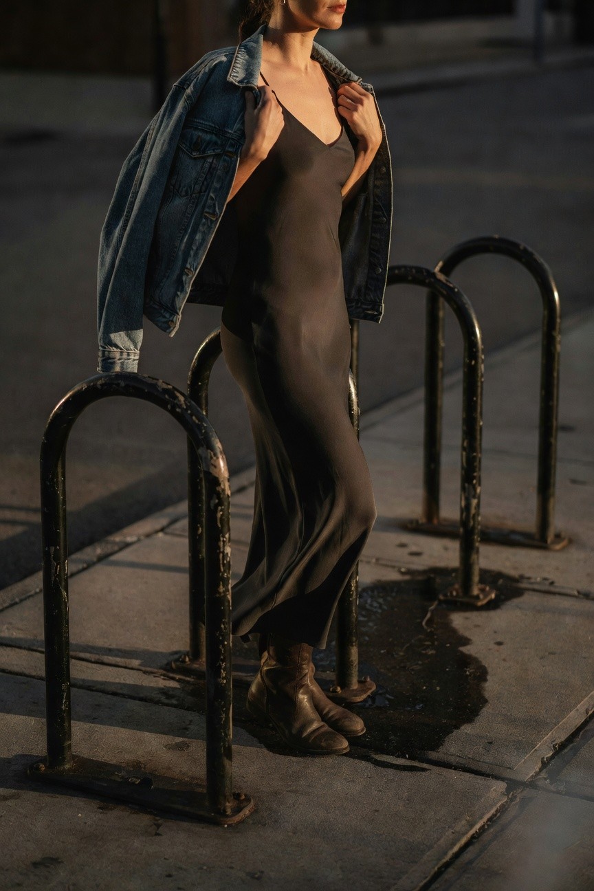 Side view of a woman in a charcoal gray spaghetti-strap slip dress layered under an open oversized denim jacket, wearing brown leather ankle boots, hands holding the jacket edges while standing by metal bike racks on a sidewalk