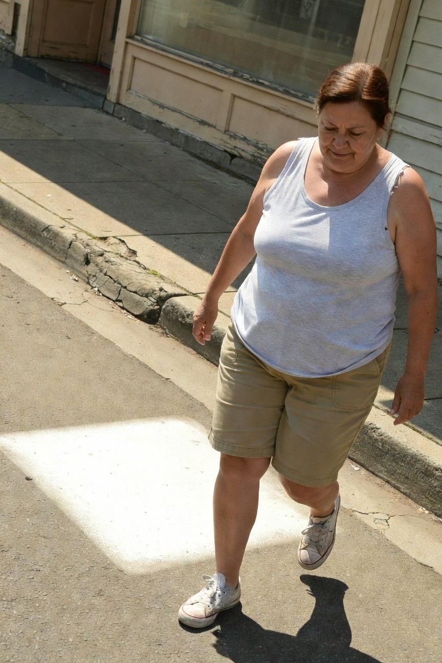 Plus-size woman over 40 in a loose gray sleeveless tank top, khaki mid-thigh shorts with pockets, and white low-top Converse sneakers, walking with hands at sides