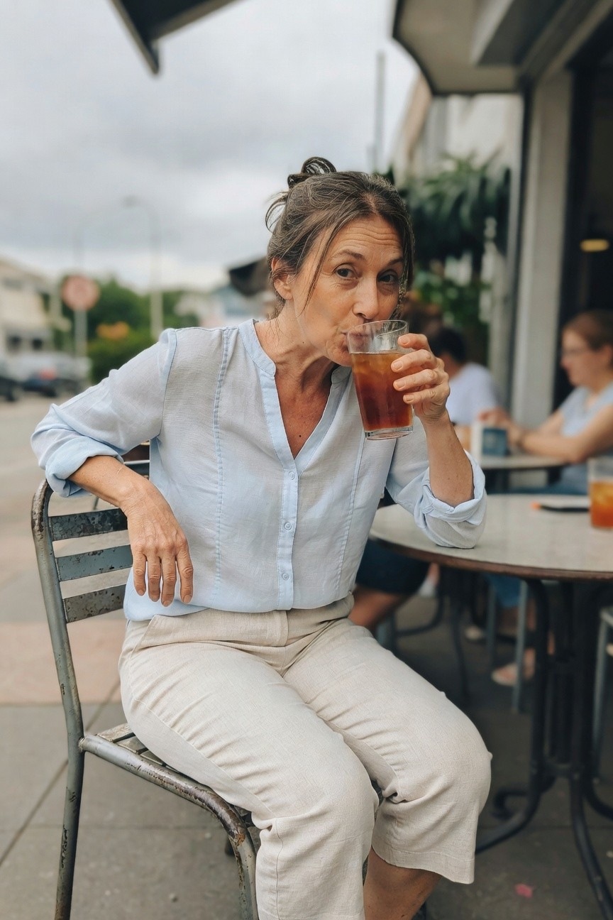 A woman over 40 in a light blue semi-sheer embroidered blouse with rolled sleeves and V-neck, paired with beige cropped linen pants, sitting at an outdoor cafe table holding an iced tea glass, relaxed posture on a metal chair.