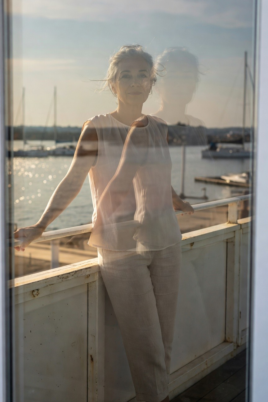 Woman over 40 in translucent peach-toned blouse and light beige linen pants leaning casually on a white railing with marina reflection in background window