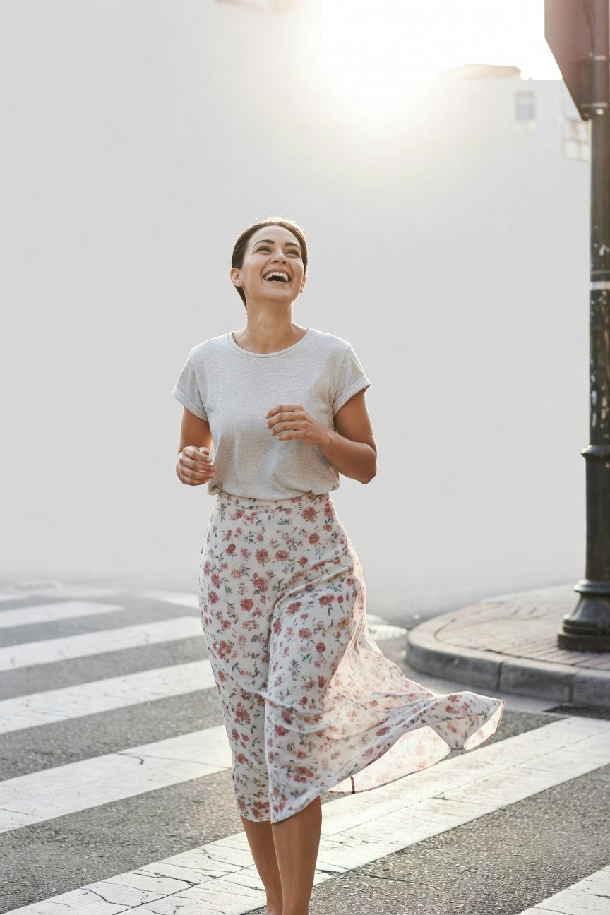 Smiling woman walking across zebra crossing in light gray short-sleeve t-shirt and sheer pink floral midi skirt with layered hem, hands in motion, urban street setting
