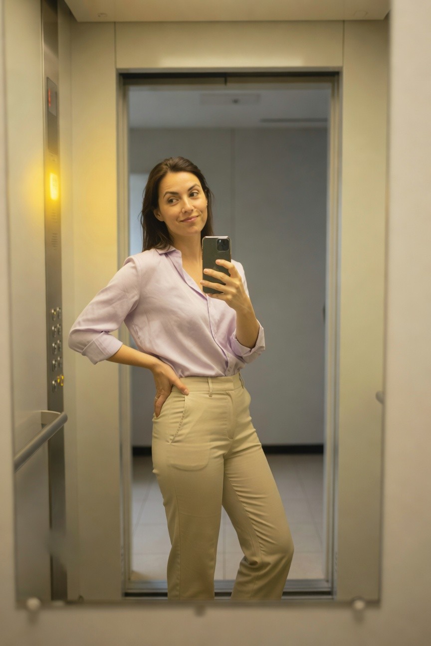 Woman taking mirror selfie in elevator wearing light purple long-sleeve button-up blouse tucked into high-waisted beige wide-leg trousers with pockets, hand on hip smiling at camera