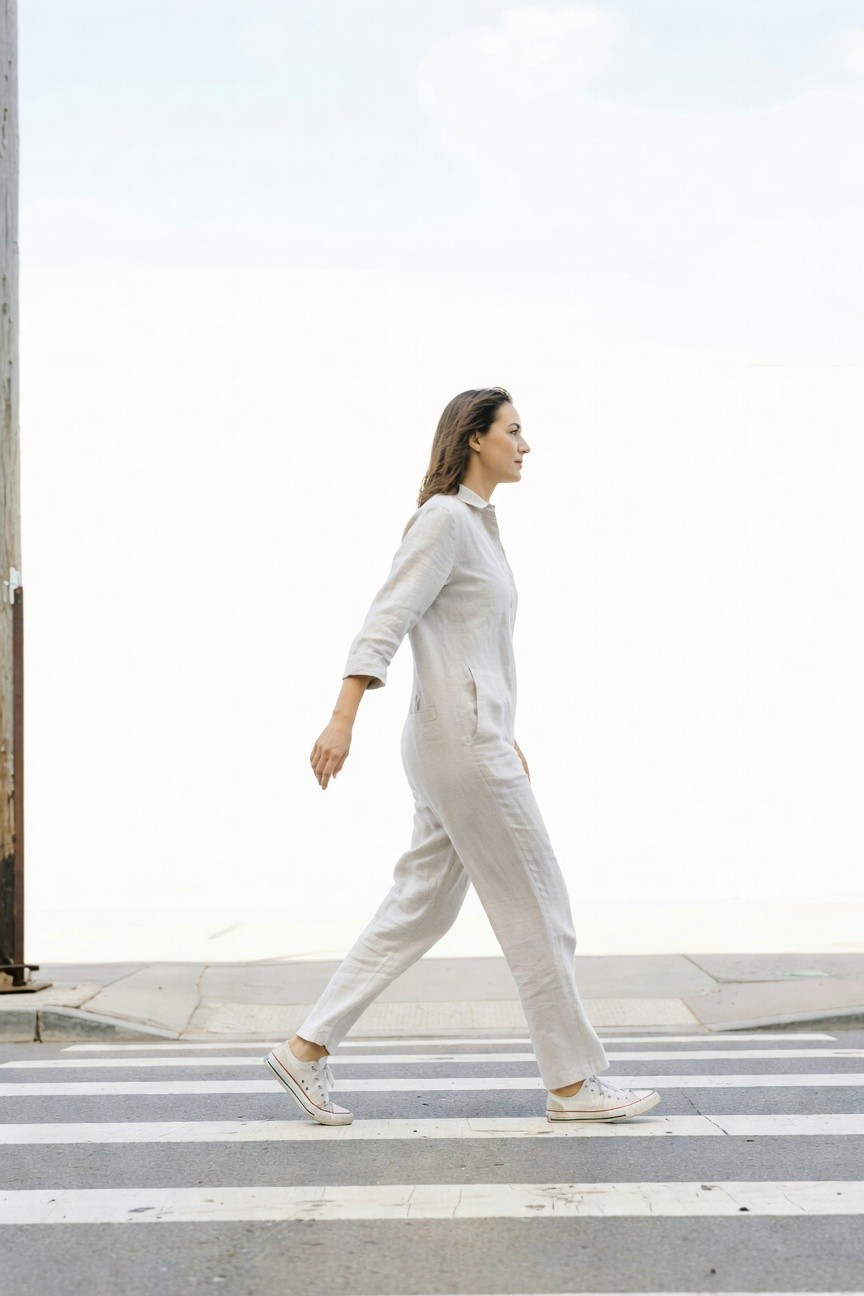 Side profile of a woman walking in a beige linen collared jumpsuit with long sleeves, wide legs, side pocket, and white Converse sneakers on a crosswalk