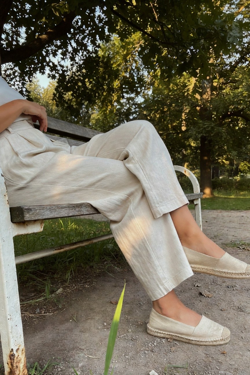Woman seated on a wooden park bench wearing cream linen wide-leg pleated pants, a loose off-shoulder white top, and matching espadrille flats with rope soles, legs extended casually