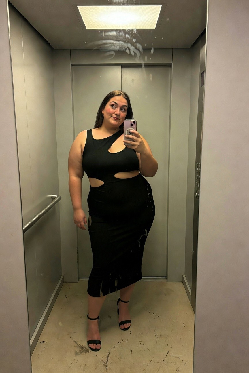 Plus-size woman taking elevator selfie in form-fitting black knit midi dress with side and midriff cutouts, fringe hem detail, and black strappy high heels, smiling at camera.