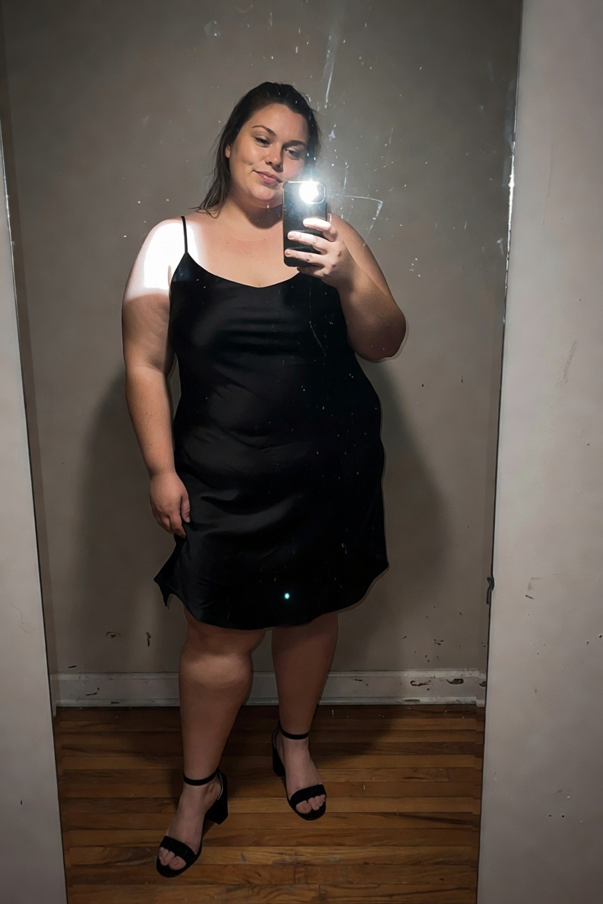 Plus-size woman in a black satin spaghetti-strap slip dress with one strap off-shoulder, mid-thigh length, paired with black strappy block heels, posing in front of a mirror selfie-style.