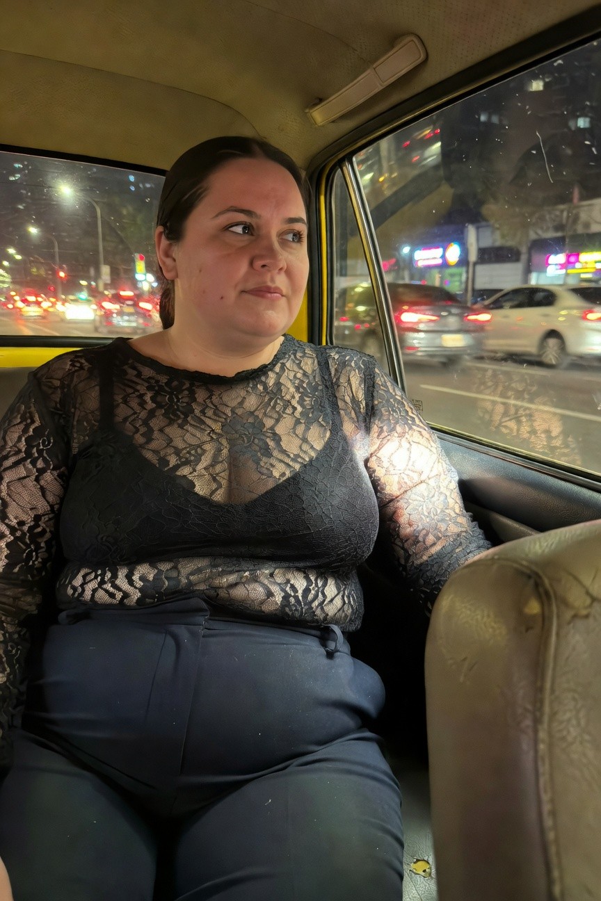 Plus-size woman in sheer black long-sleeve lace top over black bra and wide-leg black trousers, seated in yellow taxi backseat at night with city lights outside window
