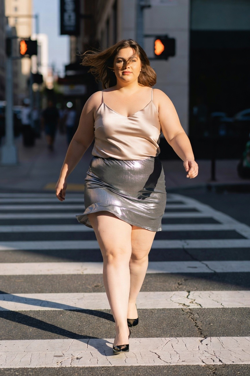 Plus-size woman wearing a beige satin camisole top with spaghetti straps, paired with a short silver metallic mini skirt and black pointed-toe high heels, walking confidently