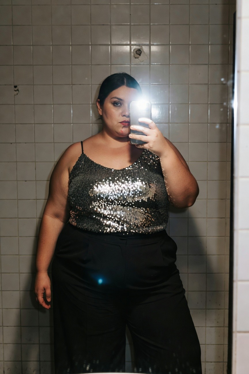Plus-size woman taking a mirror selfie in a bathroom, wearing a silver sequin spaghetti-strap top and high-waisted black wide-leg pants, dark hair parted in the middle, subtle makeup with glossy lips