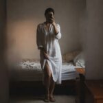 Woman taking a mirror selfie in a short white linen button-front shirt dress with long sleeves and collar, nude strap sandals, standing in bedroom lifting hem slightly on one side