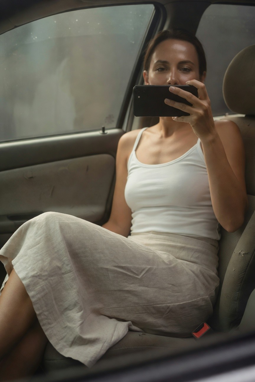 Woman seated in car passenger side wearing fitted white spaghetti-strap tank top and pale linen midi skirt with legs angled, holding phone up for selfie, seatbelt visible across lap