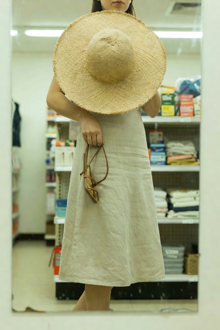 Woman in a loose beige linen midi dress holding tan leather sandals in one hand, wearing an oversized straw hat, standing barefoot in front of a store mirror reflecting shelves of products