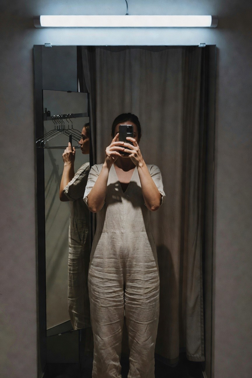 Woman taking a mirror selfie in a store fitting room while trying on a beige linen wide-leg jumpsuit with short sleeves and V-neck, holding a hanger in one hand, neutral gray curtain and clothing rack in background under fluorescent light