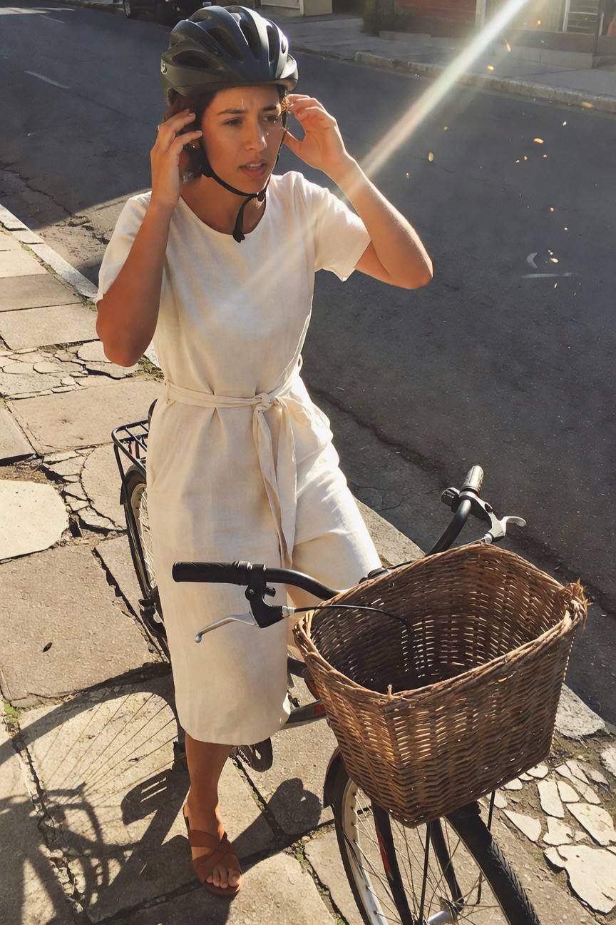 Woman standing next to a bicycle wearing a white linen midi wrap dress with belt, black bike helmet, tan cross-strap sandals, adjusting her earrings on a paved street