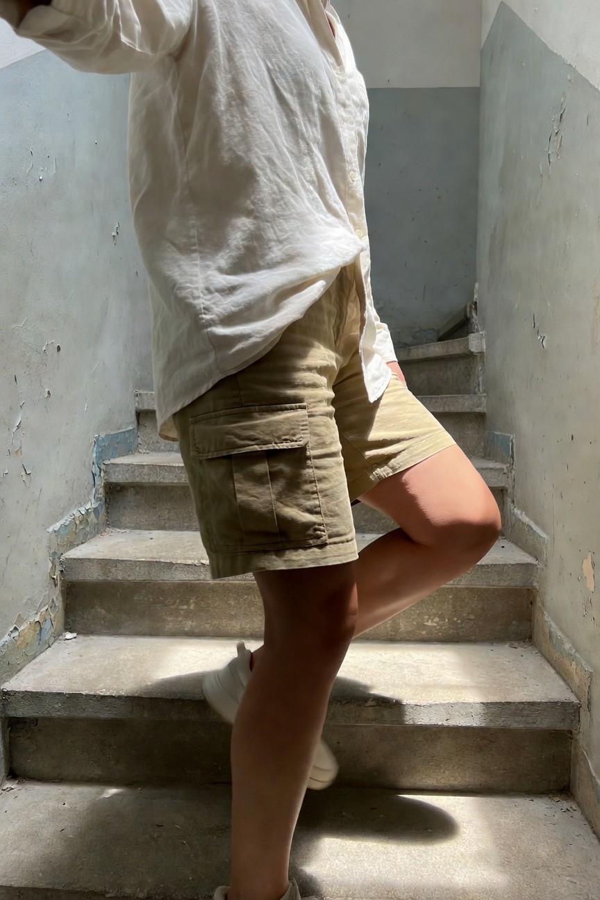 Woman standing on concrete stairs with one foot raised, wearing an oversized white linen shirt partially unbuttoned, beige khaki cargo shorts, and white sneakers, arms relaxed at sides