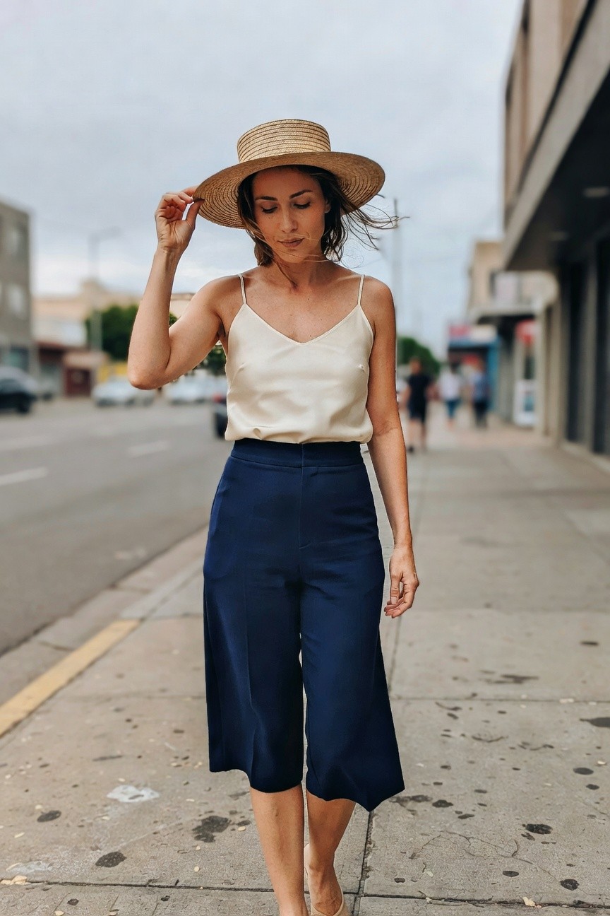 Woman walking on urban sidewalk in cream silk camisole, navy wide-leg culottes, straw hat, and nude heels, hand adjusting hat brim