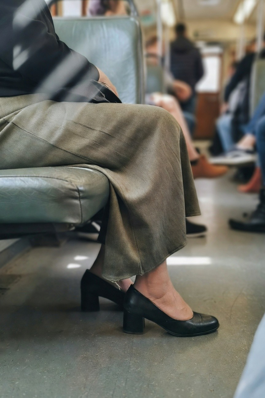 Woman seated on train bench in black top, olive wide-leg pants draped loosely, black pointed kitten heel pumps, and thin bracelet on wrist, with blurred passengers in background