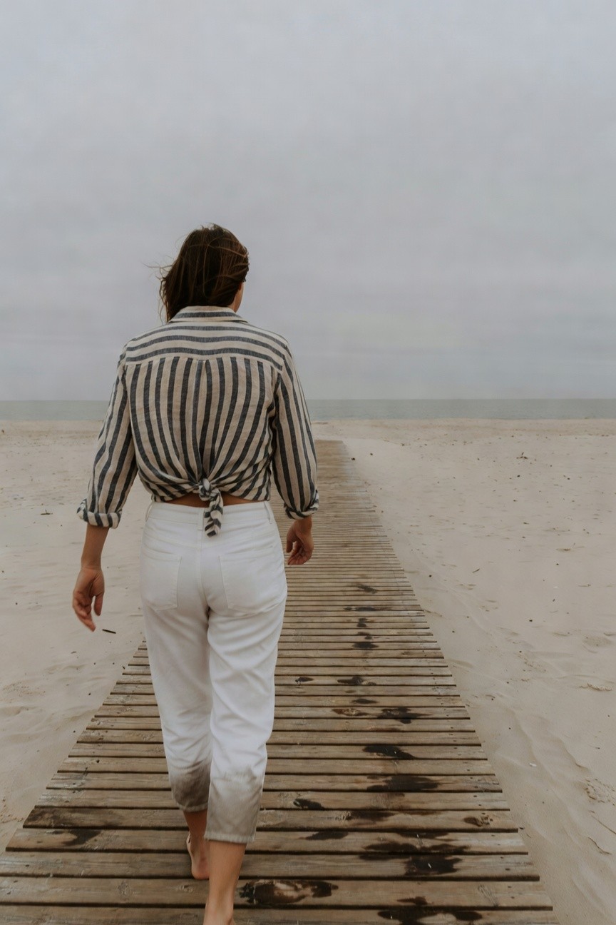 Rear view of woman walking barefoot on wooden boardwalk wearing long-sleeved striped linen shirt tied at back and white cropped straight-leg jeans