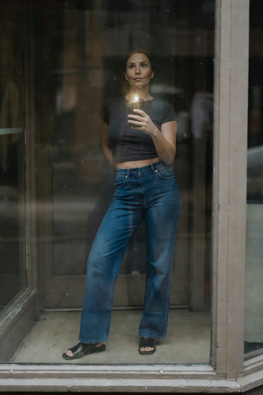 Woman standing confidently in a glass enclosure wearing a cropped gray t-shirt, high-waisted wide-leg blue jeans, and black open-toe slide sandals, holding a phone for a selfie.