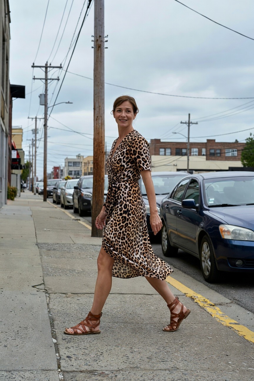 Woman striding confidently in a brown-toned leopard print wrap midi dress with v-neckline short sleeves and thigh slit paired with tan crisscross strappy flat sandals on urban sidewalk