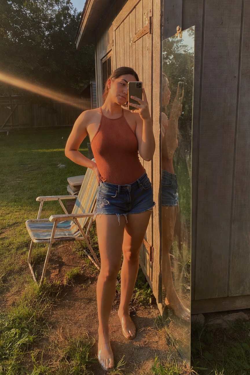 Woman in rust ribbed halter tank top and short frayed denim shorts posing confidently with hand on hip next to a wooden outbuilding mirror reflection visible clear shoes on grass
