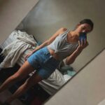 Mirror selfie of a woman with dark hair in a ponytail wearing a white cropped tank top tied at the waist, high-waisted light blue denim shorts, and tan strappy sandals, standing in a bedroom with bed in background