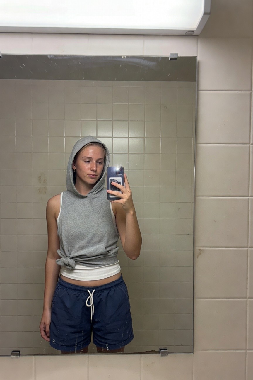 Young woman taking a mirror selfie in a tiled bathroom, wearing a gray sleeveless hoodie with hood up and waist tie exposing a white crop top underneath, paired with short navy drawstring shorts, holding her phone casually