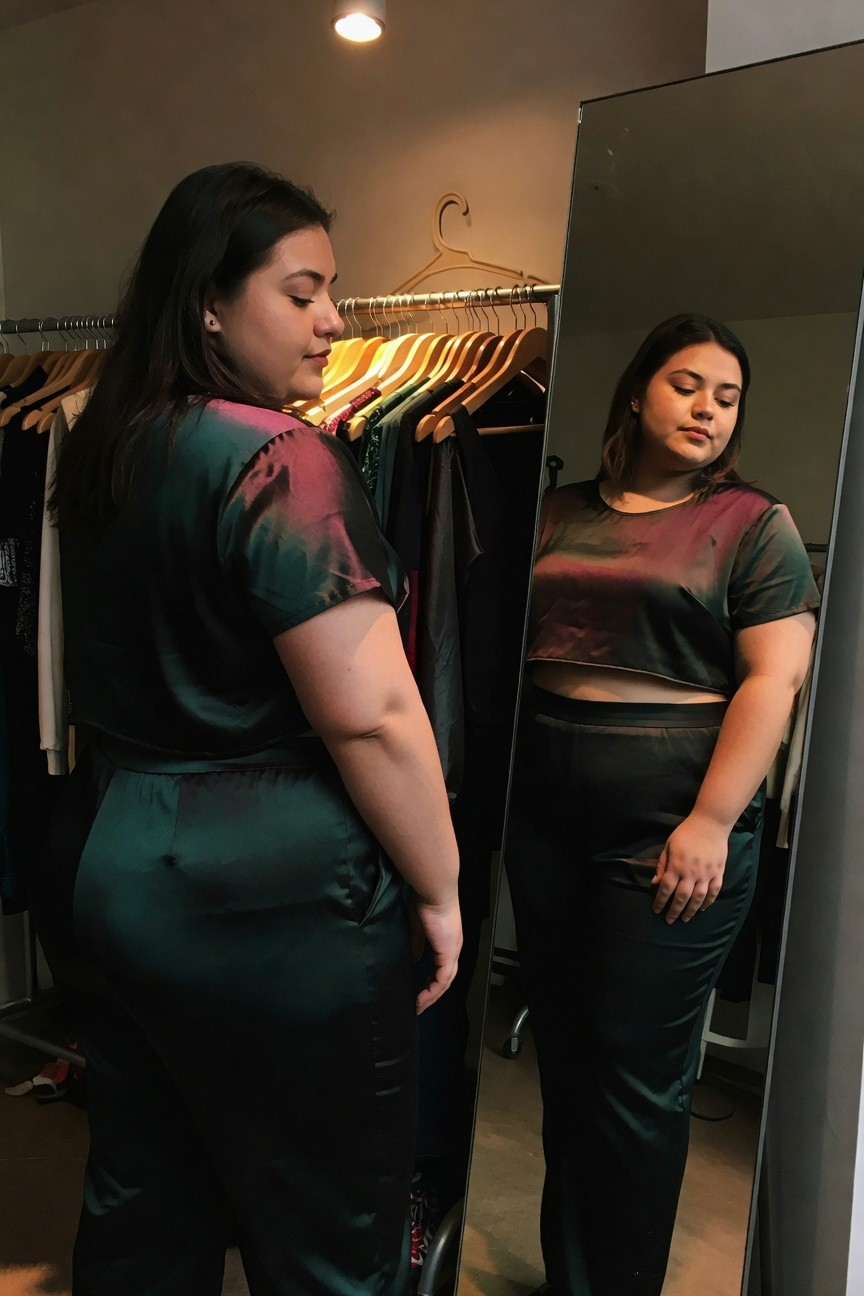Plus-size woman with dark hair posing sideways in a clothing store mirror, wearing an iridescent green-to-purple satin short-sleeve crop top and matching high-waisted wide-leg pants, racks of clothes in background