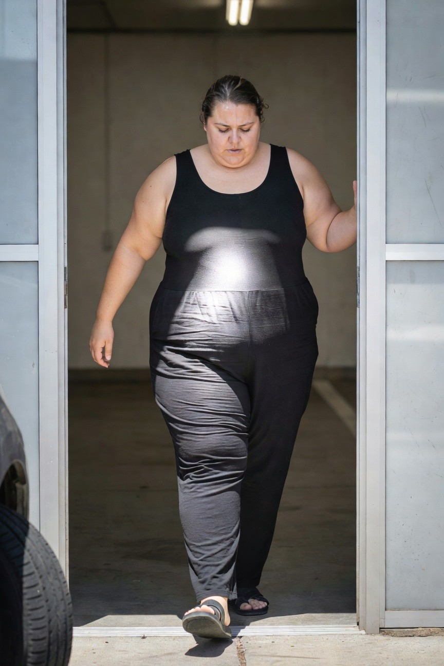 Plus-size woman walking out of a garage door wearing a sleeveless black tank-style jumpsuit with wide-leg pants and black flip-flop sandals, side view emphasizing the outfit's fit and flow
