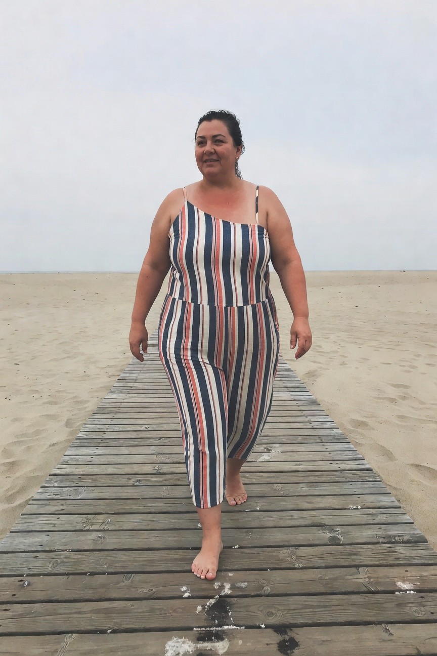 Plus-size woman with curly dark hair smiling while walking barefoot on a wooden pier in a sleeveless navy, white, and red vertically striped wide-leg jumpsuit, sand and ocean in background