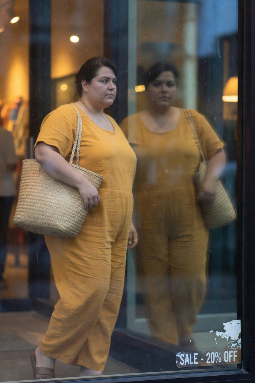 Plus-size woman in mustard yellow wide-leg linen jumpsuit, short sleeves, holding tan woven straw tote bag, wearing brown flat sandals, reflected in store window
