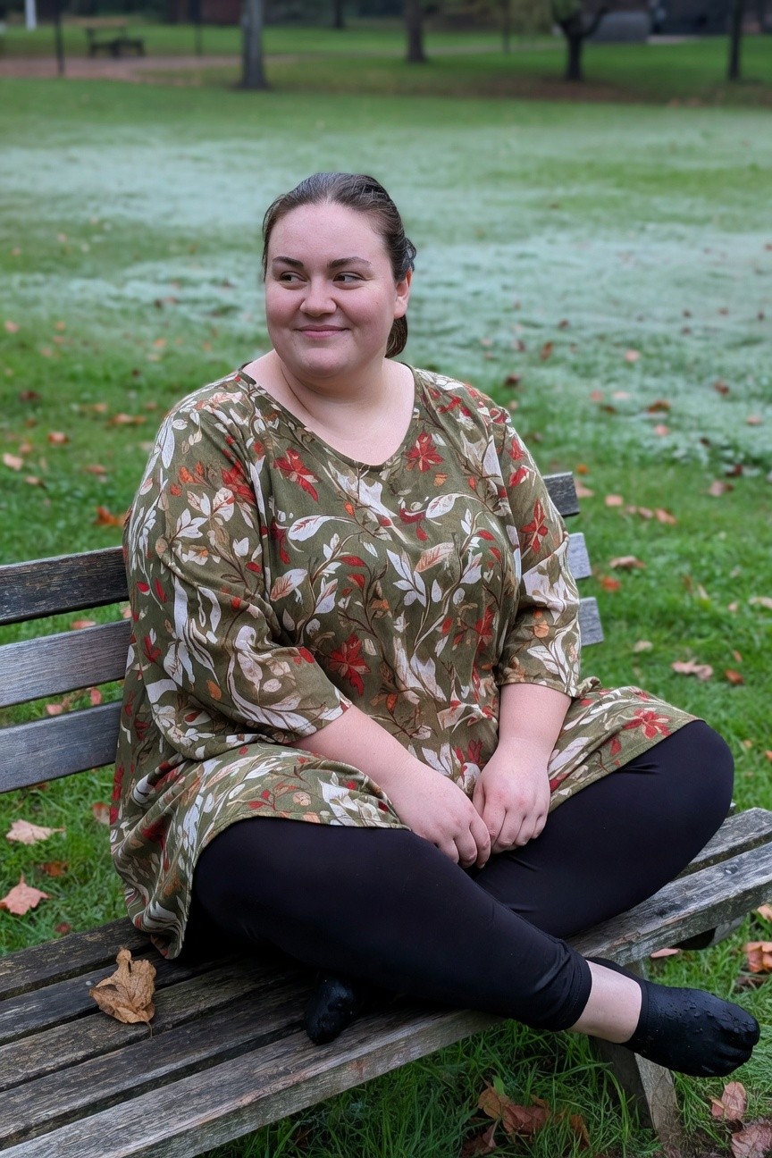 Plus-size woman sitting on a wooden bench wearing a green floral print tunic top with wide sleeves over black leggings and black toe-separating flats, smiling at the camera