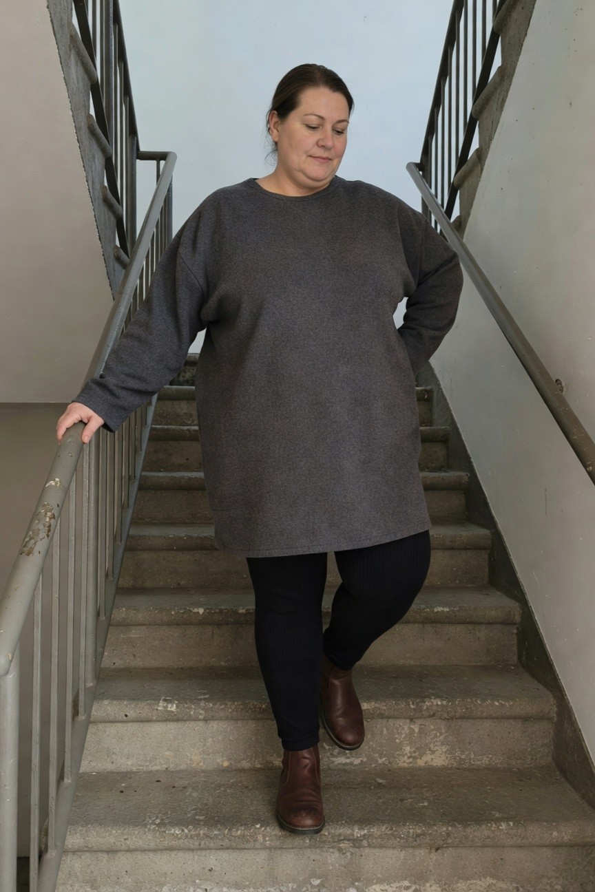Plus-size woman standing on concrete stairs in an oversized gray knit tunic dress with wide sleeves, black leggings, and brown leather ankle boots, holding a metal railing with one hand.