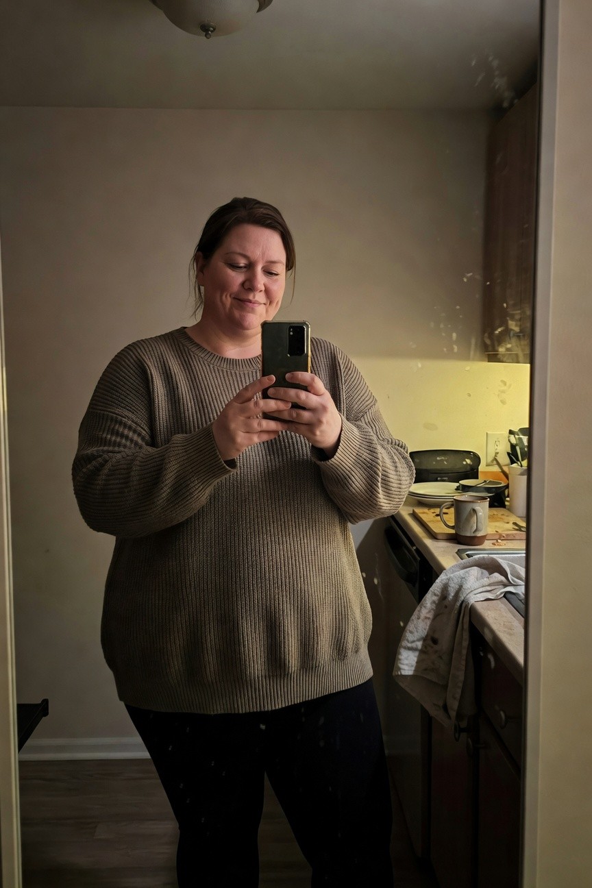Plus-size woman smiling in selfie wearing taupe chunky cable knit tunic sweater with wide sleeves and black leggings standing in front of kitchen sink mirror
