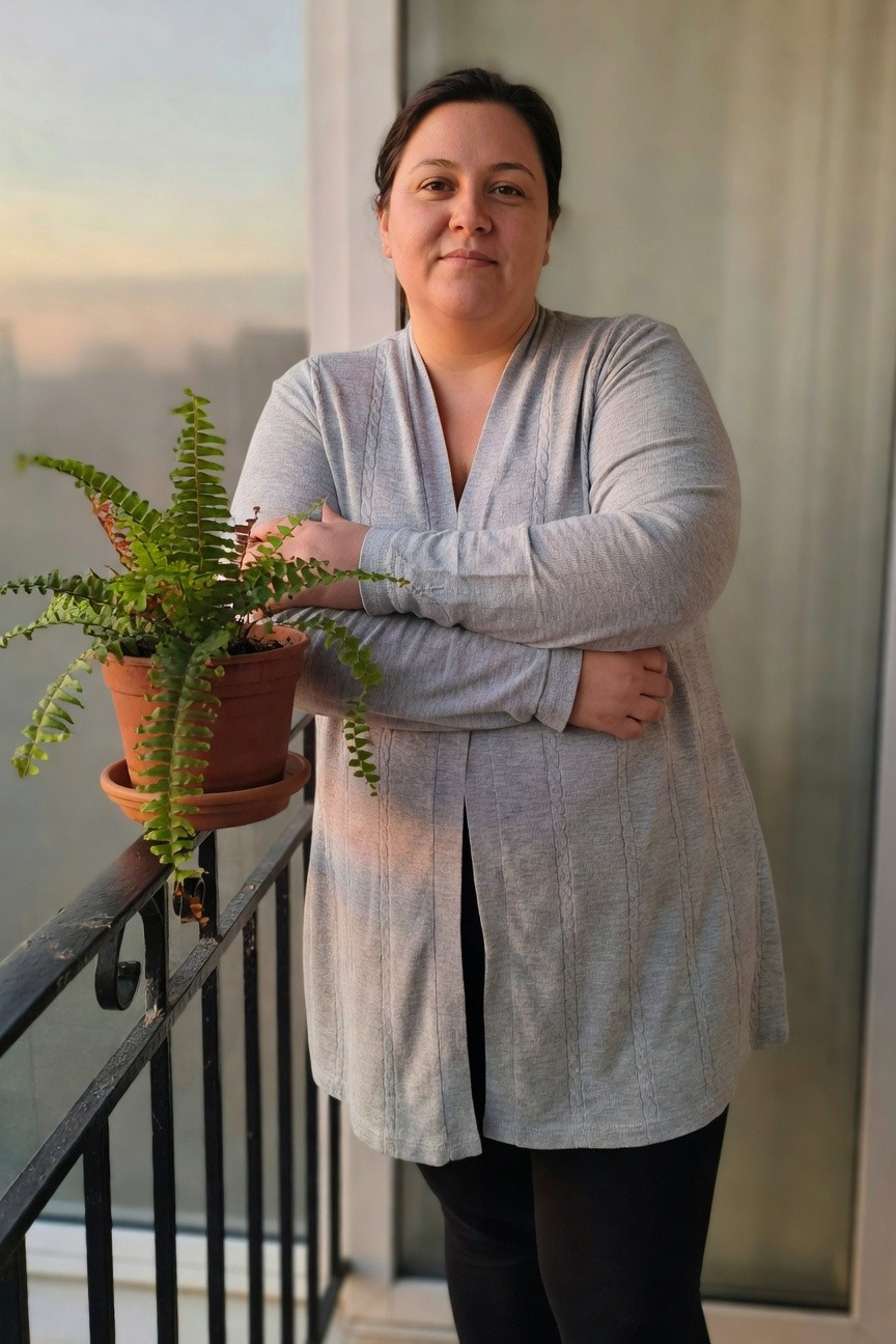 Plus-size woman in a long open-front gray knit cardigan tunic over black leggings, standing with arms crossed holding a potted fern plant on a balcony railing