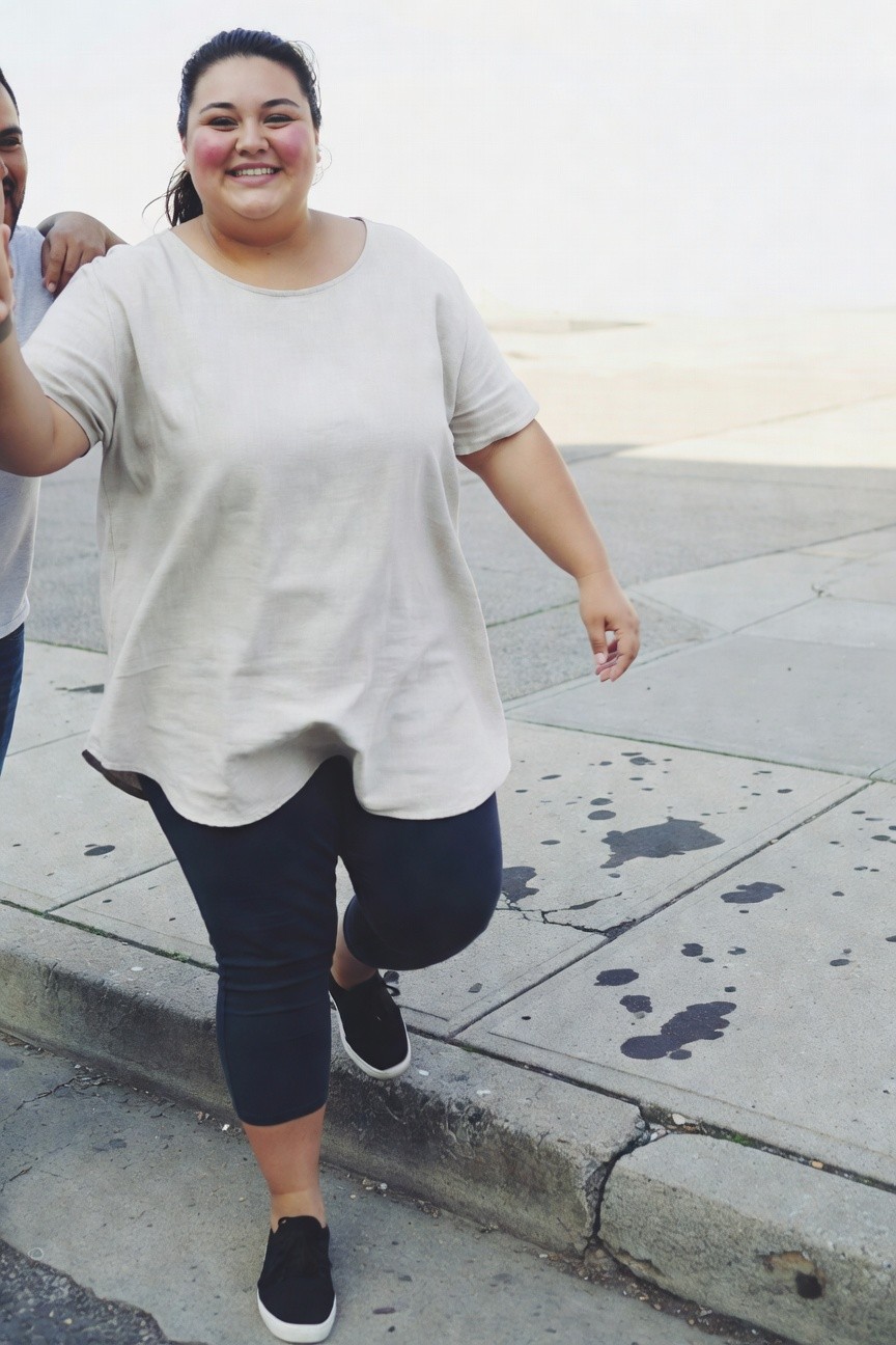 Plus-size woman smiling while walking, wearing a loose beige boat-neck tunic top, navy cropped leggings, and black sneakers, with a man's arm around her shoulder