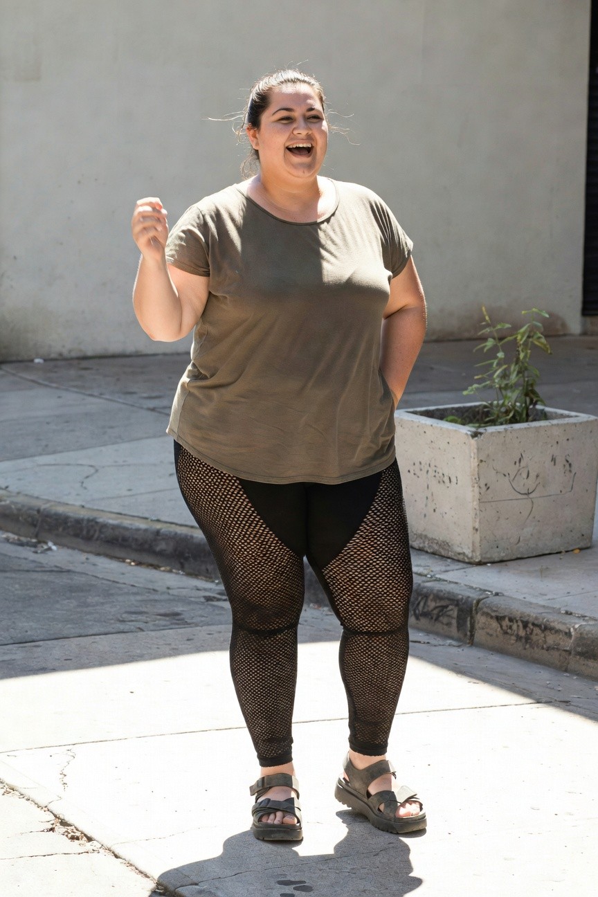 Plus-size woman laughing in an olive green loose tunic top, black mesh-panel leggings, and tan platform sandals with buckles, standing on a sidewalk