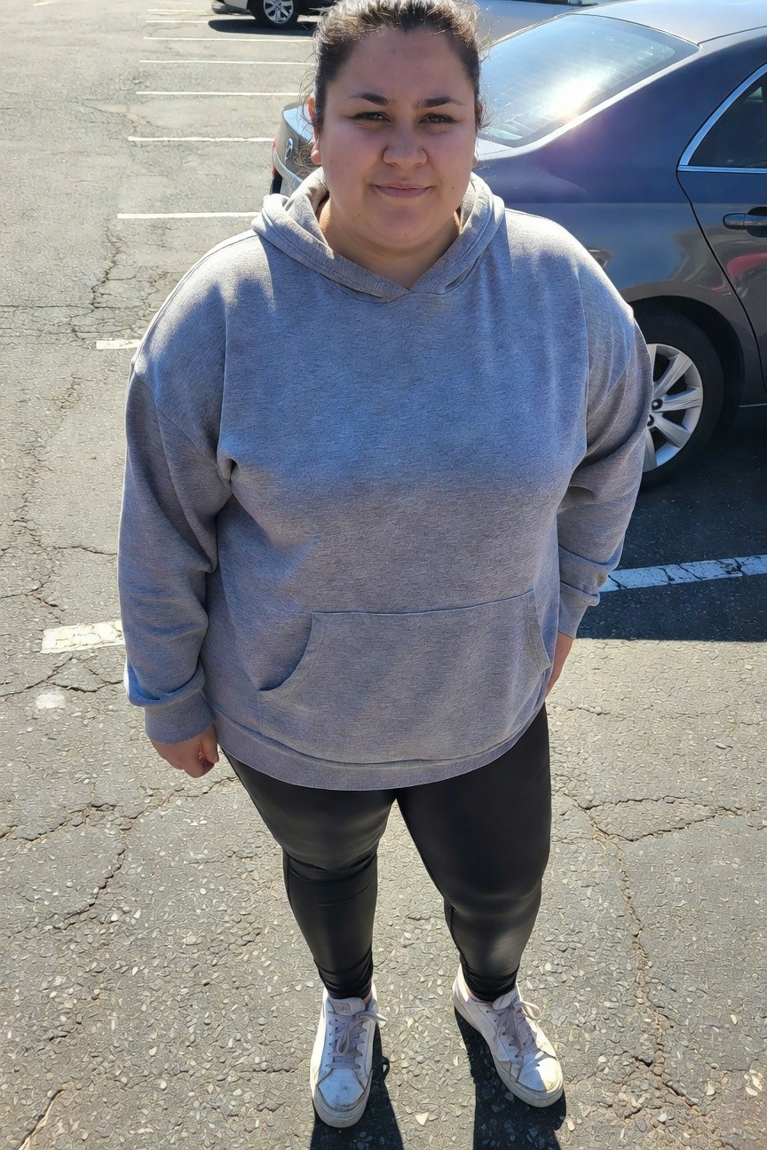 Plus-size woman standing in parking lot wearing oversized gray hoodie tunic, black leather-look leggings, and white sneakers, hands in pockets smiling at camera
