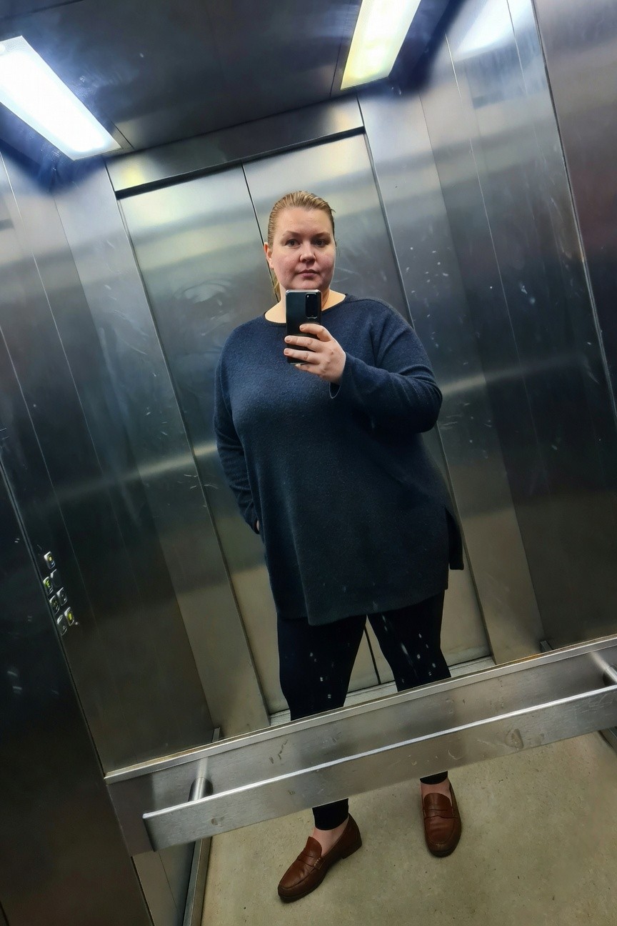 Plus-size woman with blonde hair taking a mirror selfie in a metallic elevator, wearing a loose dark navy knit tunic sweater with side slit, black splattered leggings, and brown leather loafers on a tiled floor