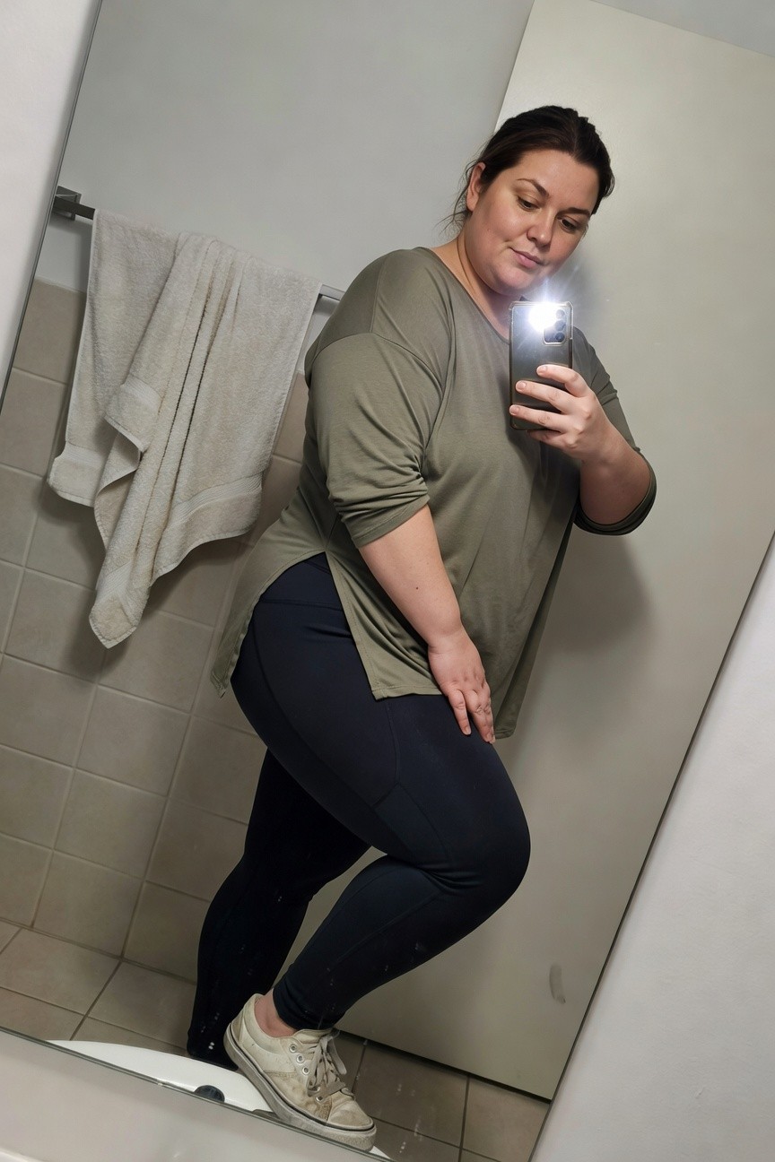 Plus-size woman in a mirror selfie wearing an oversized sage green tunic top with wide sleeves, black high-waisted leggings, and white sneakers, posing sideways in a bathroom to highlight the outfit's fit and flow.