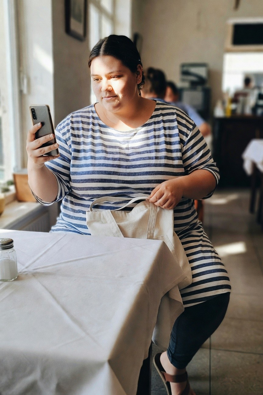 Plus-size woman sitting at a table wearing a loose navy and white horizontal striped tunic over black leggings, tan flat sandals, and a white canvas tote bag, holding a smartphone