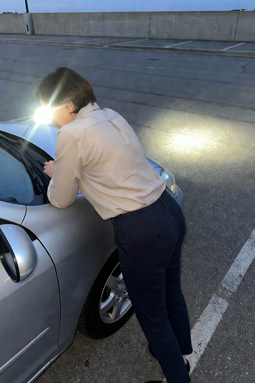 Back view of woman in light beige button-up shirt, navy blue trousers, and black flats, leaning toward a car with headlamp on forehead